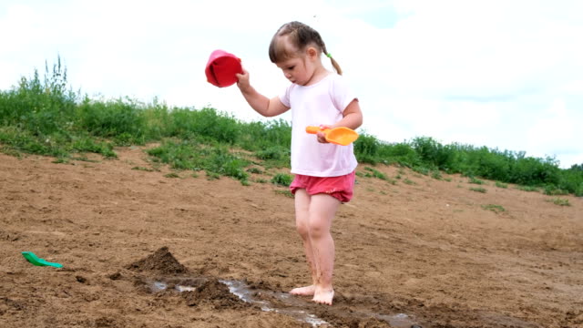 Kleines-Mädchen-gießt-Wasser-aus-einem-Eimer-in-den-Sand.-Kinder-spielen-am-Strand