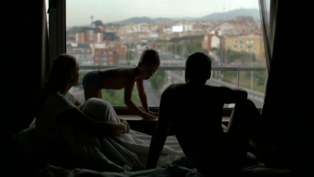 Vista-de-la-familia-en-la-cama-con-el-hijo-pequeño-contra-la-enorme-ventana-panorámica-y-paisaje-urbano,-Barcelona,-España