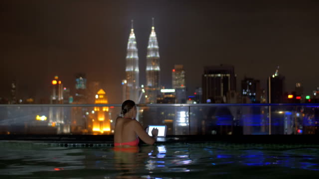 Vista-de-la-mujer-en-la-piscina-en-la-azotea-del-rascacielos-usando-tableta-contra-el-paisaje-de-la-ciudad-de-noche.-Kuala-Lumpur,-Malasia