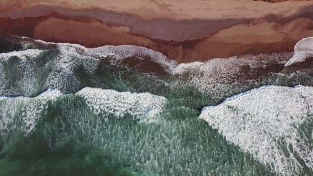 Vista-aérea-inclinación-sobre-las-olas-del-océano-y-playa-de-arena-roja