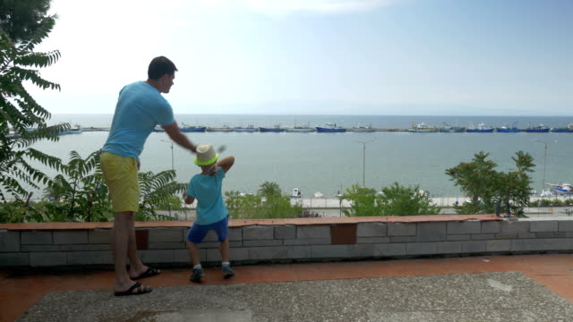 Young-man-and-son-are-throwing-rocks-against-boats-harbor-in-sea-Piraeus,-Greece