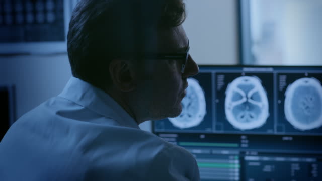 In-Control-Room-Doctor-and-Radiologist-Discuss-Diagnosis-while-Watching-Procedure-and-Monitors-Showing-Brain-Scans-Results,-In-the-Background-Patient-Undergoes-MRI-or-CT-Scan-Procedure.