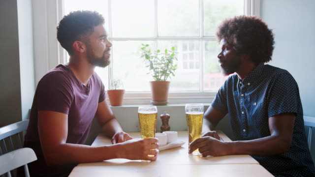 Mixed-race-young-man-and-his-black-dad-drinking-beer-and-talking-at-a-table-in-a-pub,-close-up,-side-view