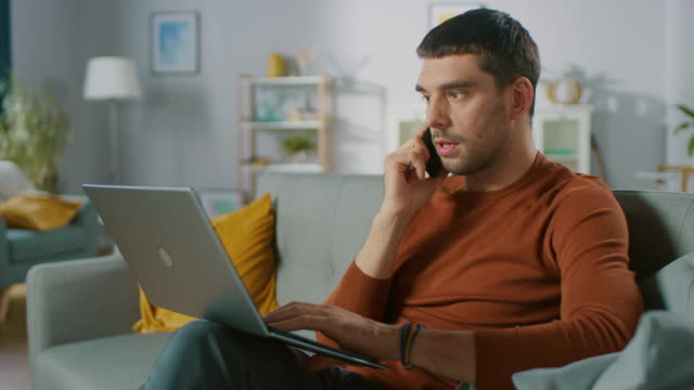 Hombre-guapo-sentado-en-el-sofá-en-casa,-habla-en-el-teléfono-móvil-y-utiliza-el-ordenador-portátil.