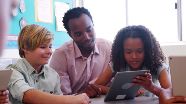 Male-teacher-helping-two-young-kids-using-tablet-in-class