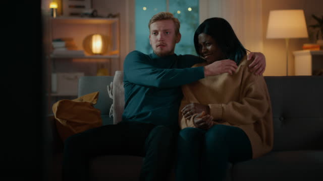 Happy-Young-Couple-Watching-Horror-Movie-on-TV-while-Sitting-on-a-Couch,-they-got-Scared,-Girl-Covers-eyes.-Handsome-Caucasian-Boy-and-Black-Girl-in-Love-Spending-Time-Together.-Moving-Camera-Shot.