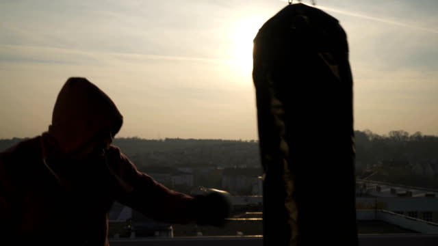 Boxer-with-fighting-gloves-stands-in-guard-punches-bag-in-sunset,-close-up,-city,-sportsman-practicing,-power-training,-strong-guy-hard-exercising,-strength-exercises,-workout,-handheld,-sunny-day.