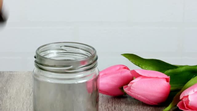 A-woman-is-taking-a-coin-in-a-jar,-which-is-placed-on-a-wooden-table-and-has-a-pink-tulip