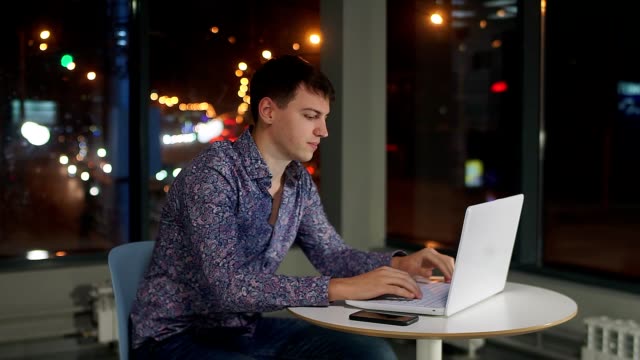 Un-hombre-escribiendo-en-un-teclado-de-ordenador-portátil,-en-la-oficina-en-una-gran-ventana,-contra-la-ciudad-de-noche.