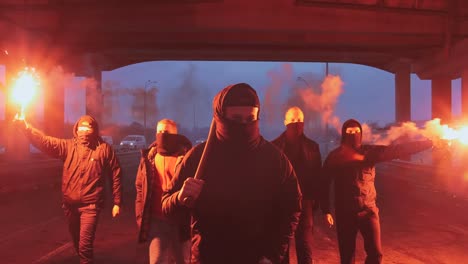 Group-of-young-men-in-balaclavas-with-red-burning-signal-flare-walking-on-the-road-under-the-bridge,-slow-motion