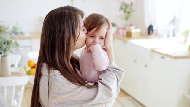 Pflegende-junge-Mutter-beruhigend,-küsst-und-streichelt-ihre-niedliche-kleine-Tochter,-die-zu-Hause-bitterlich-in-ihren-Armen-weint