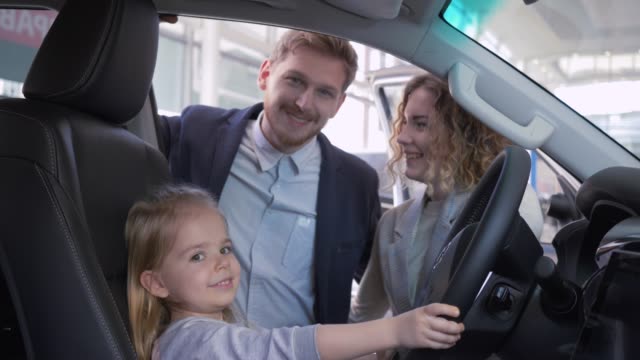 happy-childhood,-sweet-kid-girl-behind-wheel-of-automobile-together-with-mother-and-father-while-buying-family-machine-at-sales-center-close-up