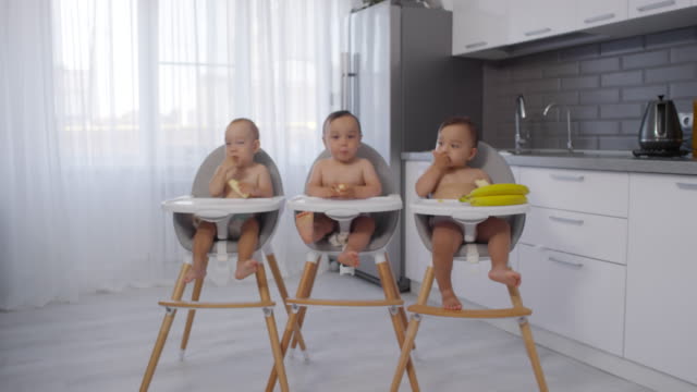 Asiatische-Baby-Triplets-sitzen-in-Hochstühlen-in-der-Küche-und-essen-Bananen