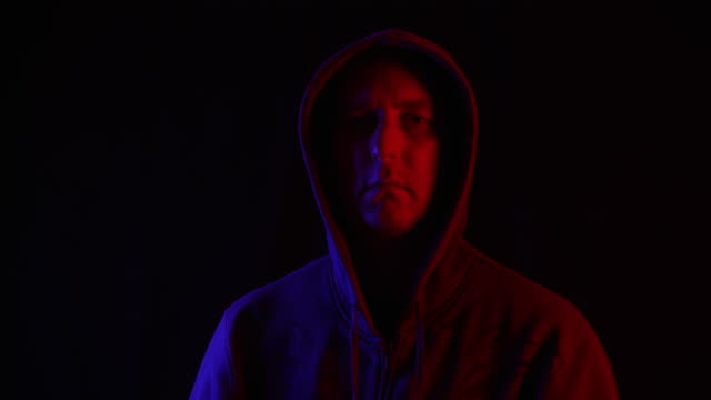 Man-in-dark-hoodie-looking-up-to-camera-on-black-background-in-blue-and-red-lighting.-Portrait-mysterious-man-in-hood-in-dark-studio-with-red-and-blue-backlight.