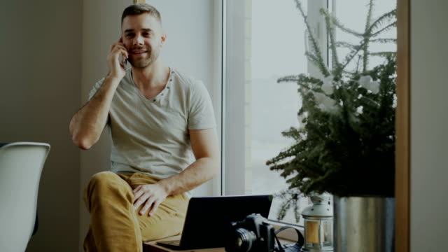 Attraktive-junge-Mann-sprechen-Telefon-sitzt-auf-der-Fensterbank-mit-Laptop-und-Kamera-zu-Hause
