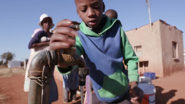Close-up-of-Young-african-boy-drinking-water-from-a-tap-while-woman-line-up-to-collect-water-in-plastic-containers-due-to-severe-drought-in-South-Africa