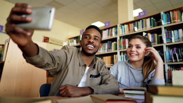 Africanos-merican-y-caucásico-se-divierten-sonriendo-y-tomando-fotos-selfie-en-cámara-del-smartphone-en-la-biblioteca-de-la-Universidad.-Los-estudiantes-alegres-tienen-descanso-mientras-preparan-para-examen