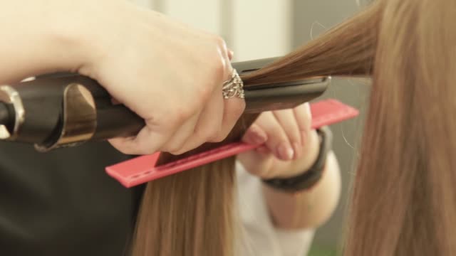 Friseur-mit-Zange-Haar-und-Kamm-für-Haarglättung-Art-und-Weisemodell-im-Beauty-Salon.-Friseur-richten-Sie-lange-Haare-während-Frau-Haarstyling-im-Friseur-Salon-hautnah