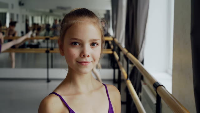 Primer-plano-retrato-de-hermosa-niña-en-mono-en-clase-de-ballet,-sonriendo-y-mirando-a-cámara.-Otros-alumnos-están-haciendo-ejercicios-de-fondo.