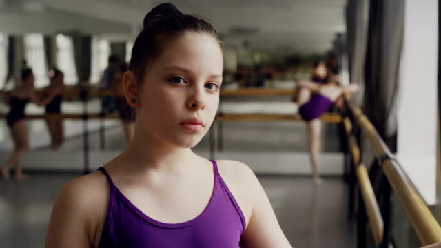 Close-up-portrait-of-dark-haired-little-girl-in-bodysuit-standing-in-ballet-class-and-looking-at-camera.-Other-students-are-practising-in-background.