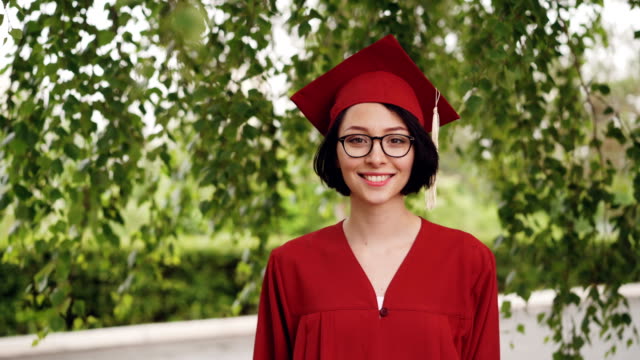 Portrait-of-joyful-young-woman-graduating-student-in-gown-and-mortar-board-smiling-and-looking-at-camera-standing-under-the-tree-on-campus.-Youth-and-education-concept.
