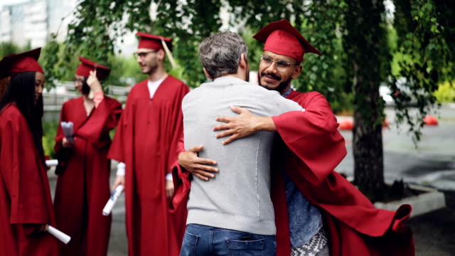 Proud-teacher-is-congratulating-student,-shaking-his-hand-and-hugging-him-on-graduation-day-standing-on-campus-in-traditional-grad-garment.-Education-and-people-concept.