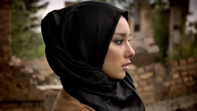 Joven-musulmana-hijab-mirando-lejos-y-al-cámara,-preocupada-y-con-miedo,-arruinado-edificio-de-fondo