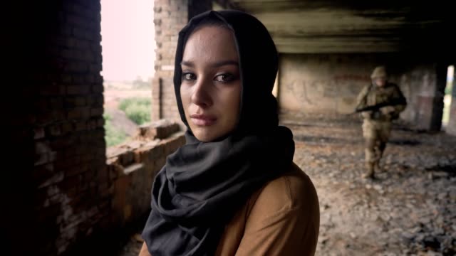 Joven-musulmana-hijab-llorando-y-mirando-a-cámara,-soldado-con-el-arma-en-el-fondo,-abandonado-edificio,-concepto-de-terrorismo