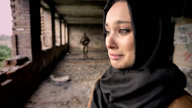 Joven-mujer-musulmana-triste-en-hijab-llorando-cuando-armado-a-soldado-va-hacia-la-mujer,-abandonada-del-edificio,-concepto-de-guerra