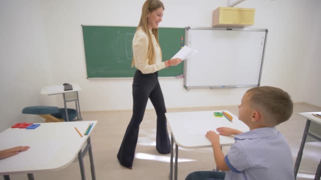 prueba-de-la-escuela,-sonriendo-a-profesor-femenino-distribuye-hojas-blancas-de-papel-para-comprobar-conocimientos-de-los-escolares-en-el-escritorio-durante-la-lección-en-clase-en-la-escuela