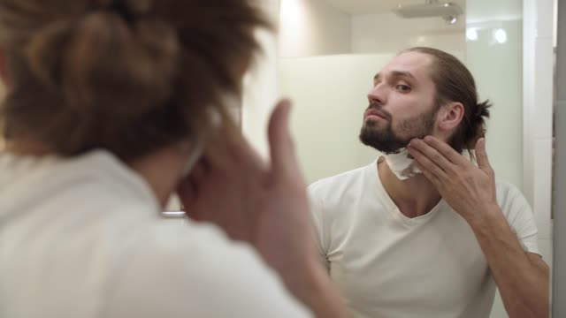 Aplicación-de-espuma-de-afeitar-sobre-la-piel-de-la-cara-y-mirando-en-el-espejo-del-hombre