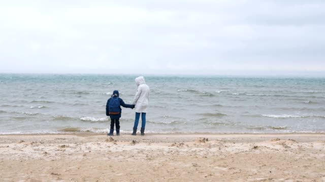 Mamá-e-hijo-están-caminando-en-la-playa-de-arena-del-mar-en-invierno,-vista-posterior.