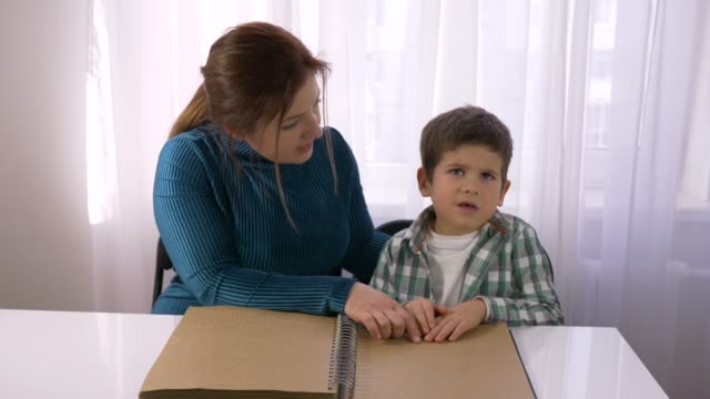Schwierige-Schulbildung-für-blinde-Kinder,-junge-Lehrerin-lehrt-sehbehinderte-Junge,-Braille-Bücher-mit-Symbolen-Schrift-sitzen-am-Tisch-im-Zimmer