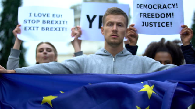 Young-man-holds-European-Union-flag,-activists-with-freedom-slogans-on-backdrop
