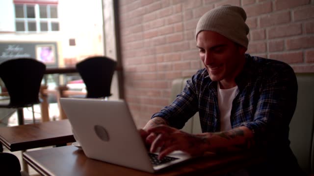 Young-hipster-chatting-online-using-laptop,-laughing-at-coffee-shop