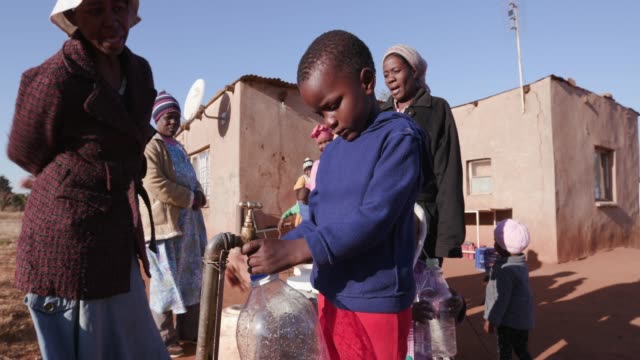 Young-african-boy-collecting-water-from-a-tap-while-woman-line-up-to-collect-water-in-plastic-containers-due-to-severe-drought-in-South-Africa