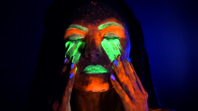 Portrait-of-girl-smears-and-rubs-fluorescent-paint-under-eyes.-Dye-glowing-near-UV-black-light.-Woman-with-braids-in-neon-light.-Night-club,-party,-halloween-psychedelic-concepts