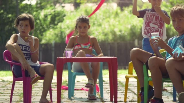 Junge-glückliche-Kinder-im-Sommerlager-sitzen-bunte-Kunststoff-Stühle-Trinken-Essen-Snacks-Spaß-außerhalb-im-grünen-Rasen-Feld-Kinder-Partei-Zeitlupe