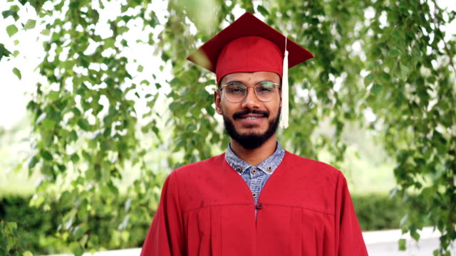Portrait-of-bearded-mixed-race-man-graduating-student-in-gown-and-mortar-board-smiling-and-looking-at-camera-standing-outdoors-on-campus.-People-and-education-concept.