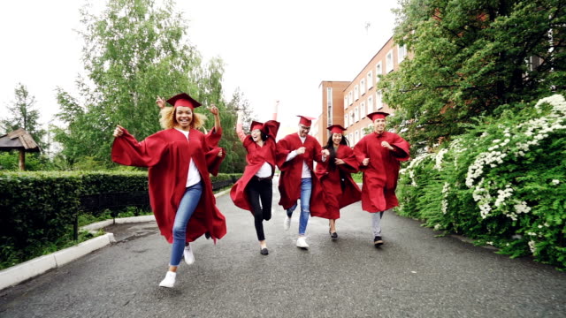 Dolly-Schuss-von-aufgeregt-Absolventen-auf-dem-Campus-tragen-Kleider-und-Trachtenhüte-feiert-Ende-des-Studiums.-Höhere-Bildung,-Jugend-und-Glück-Konzept.