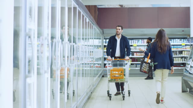 En-el-supermercado:-guapo-empuja-compras-la-tarjeta-y-navega-por-los-productos-en-la-sección-de-congelados.-Hombre-busca-en-refrigerador-de-puerta-de-cristal,-elegir-productos-lácteos.-Otro-cliente-comercial.