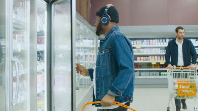 Im-Supermarkt:-stilvolle-Afroamerikaner-Kerl-mit-Kopfhörer-wählt-Produkte-in-die-gefrorene-Ware-aus-dem-Kühlschrank-und-setzt-sie-in-den-Warenkorb.-Slow-Motion.