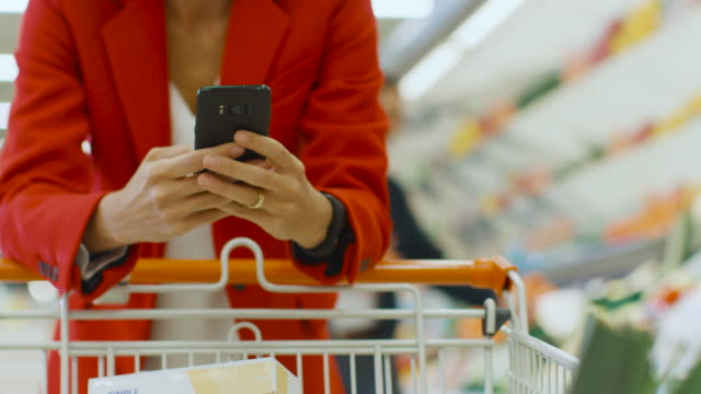 At-the-Supermarket:-Woman-Uses-Smartphone,-Leans-on-the-Shopping-Cart.-In-the-Big-Mall-Woman-Browsing-In-Internet-on-Her-Mobile-Phone.-Focus-on-Hands-Holding-Mobile-Phone.