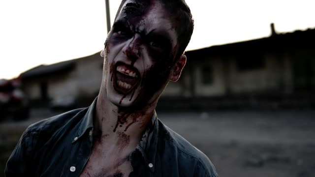 Portrait-of-a-male-zombie-with-bloody-teeth-and-wounded-face-screaming-and-shouting.-Halloween,-filming,-staging-concept.-Blurred-abandoned-town-on-the-background