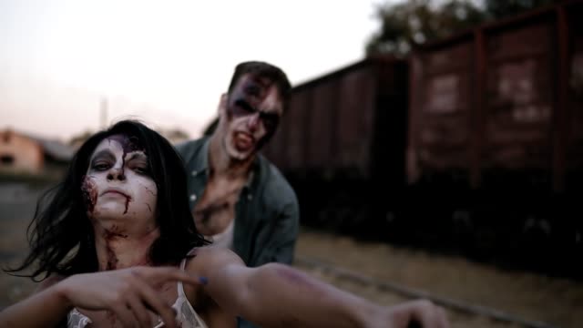 Portrait-of-a-female-zombie-with-wounded-face-in-bloody-dress-shouting-and-zombie-man-behind-her-coming-on-.-Abandoned-town-and-railway-wagons-on-the-background