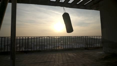 Symbolischen-Schuss-mit-Silhouette-der-Boxsack-beim-schwingen-auf-Terrasse-mit-Zaun-im-Sonnenuntergang,-Blick-auf-Stadt,-Konzept-Sport,-üben,-Ausbildung,-Training,-Übungen.-Handheld,-sonnigen-Tag-im-Freien.