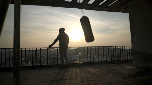 Boxing-bag-swinging,-man-comes-on-terrace-and-looks-at-city,-sunset,-silhouettes,-sportsman-practicing,-power-training,-strong-guy-hard-exercising,-strength-exercises,-workout,-handheld,-sunny-day.