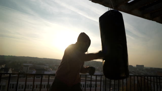 Boxer-performs-series-of-punching-in-boxing-bag-during-power-training-in-sunset,-sportsman-practicing,-power-training,-strong-guy-hard-exercising,-strength-exercises,-workout,-handheld,-sunny-day.