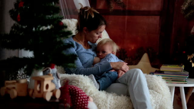 Mutter-stillen-ihren-kleinen-Sohn-sitzen-im-gemütlichen-Sessel-in-der-Nähe-von-Weihnachtsbaum,-Winter