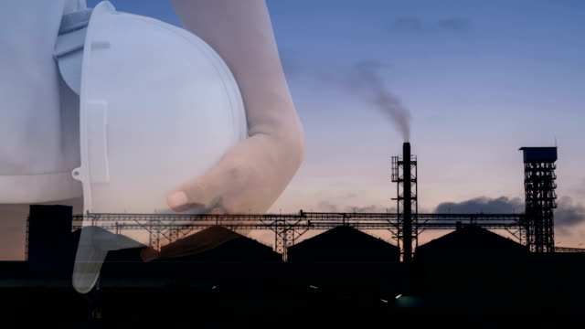Double-exposure.-Engineer-or-worker-hold-helmet.-Background--factory-industry-Set-of-storage-tanks-raw-material-feed-mills.-Safety-at-work-concept.-Time-Lapse
